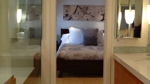 Ein modernes Hotelzimmer mit einer Glaswand, die Bad und Schlafbereich voneinander trennt. Das Zimmer bietet ein Bett mit weißer und grauer Bettwäsche, einen großen Spiegel und ein an der Wand befestigtes Kunstwerk.