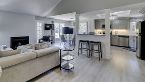 A modern living room with a sofa, a TV, and a fireplace. There is a kitchen area with a fridge, stove, and bar stools. - Sunset Rentals | Evian 233 (Hilton Head Island)