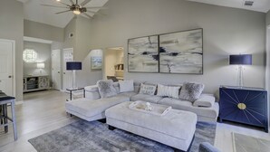 A modern living room with a sectional sofa, a coffee table, and a blue cabinet. There is a ceiling fan and abstract wall art. - Sunset Rentals | Evian 233 (Hilton Head Island)