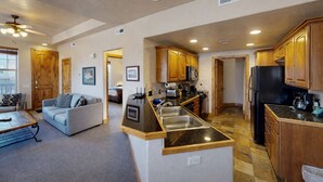 A spacious living area with a large sofa, a coffee table, and a dining table. The kitchen features wooden cabinets, a black refrigerator, and a granite countertop. - Foxpoint 2 Bedroom Condo by RedAwning (Park City)