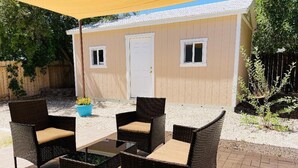 Een terras buiten met rieten meubilair en een kleine tafel, een beige gebouw met een witte deur, en een houten hek op de achtergrond.