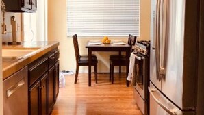 Een keuken met houten kastjes, een eettafel met stoelen, een koelkast en een raam met jaloezieën.