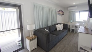 A modern living room with a grey sofa, a small white side table, and a dining area with a white table and chairs. - UNIT   1.. OPPOSITE THE OCEAN.. BEACH ..RIVER..PET FRIENDLY. 1 FREE PARKING  
 
 (HARRINGTON)