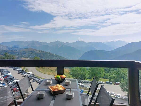 A table with a bowl of fruit, a cup, and a tray of pastries, overlooking a parking area and mountains. - Les Terrasses du Corbier by Interhome (Le Corbier)
