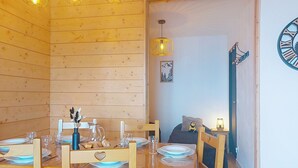 A wooden interior with a dining table set for four, a bed with a nightstand, and a wall clock. - Les Terrasses du Corbier by Interhome (Le Corbier)