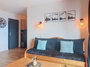 A cozy living room with a dark sofa, two light blue cushions, a wooden coffee table with a vase and remote control, and wall-mounted picture frames depicting mountain landscapes. - Les Terrasses du Corbier by Interhome (Le Corbier)
