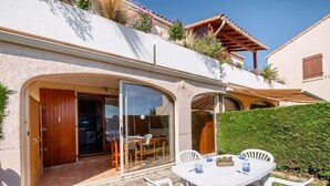 Ein Haus mit einer Terrassenanlage, inklusive Tisch und Stühlen, einem Glastür-Eingang und einem kleinen Garten mit einer Hecke.
