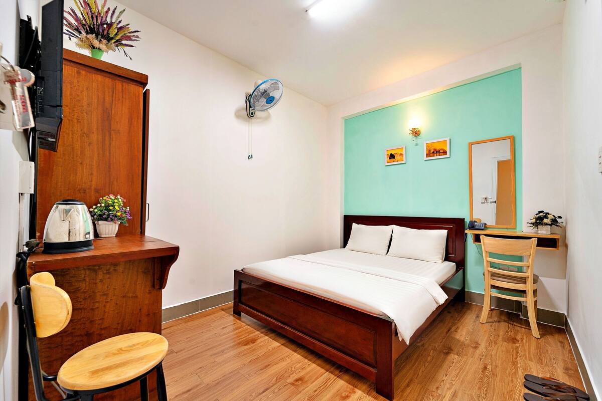 A bedroom with a wooden bed, a desk with a chair, a small table with a kettle, and a wall-mounted fan.