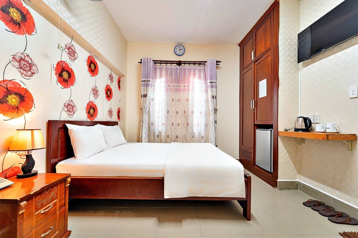 A bedroom with a bed, a nightstand, a dresser, a TV, a refrigerator, and a shelf with cups and a kettle.