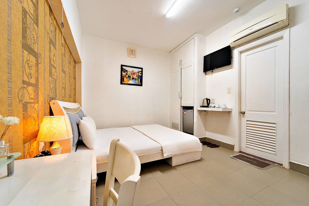 A small, clean room with a bed, a desk with a lamp, a refrigerator, and a door with a vent.