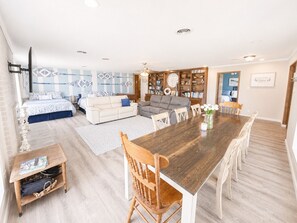A spacious living area with a large wooden dining table, a sofa set, a bookshelf, and a decorative wall clock.