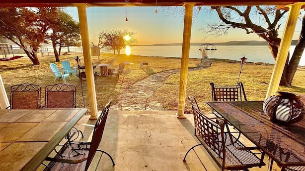 Uma área de estar coberta ao ar livre com mesas e cadeiras, com vista para uma praia, um cais e o pôr do sol.