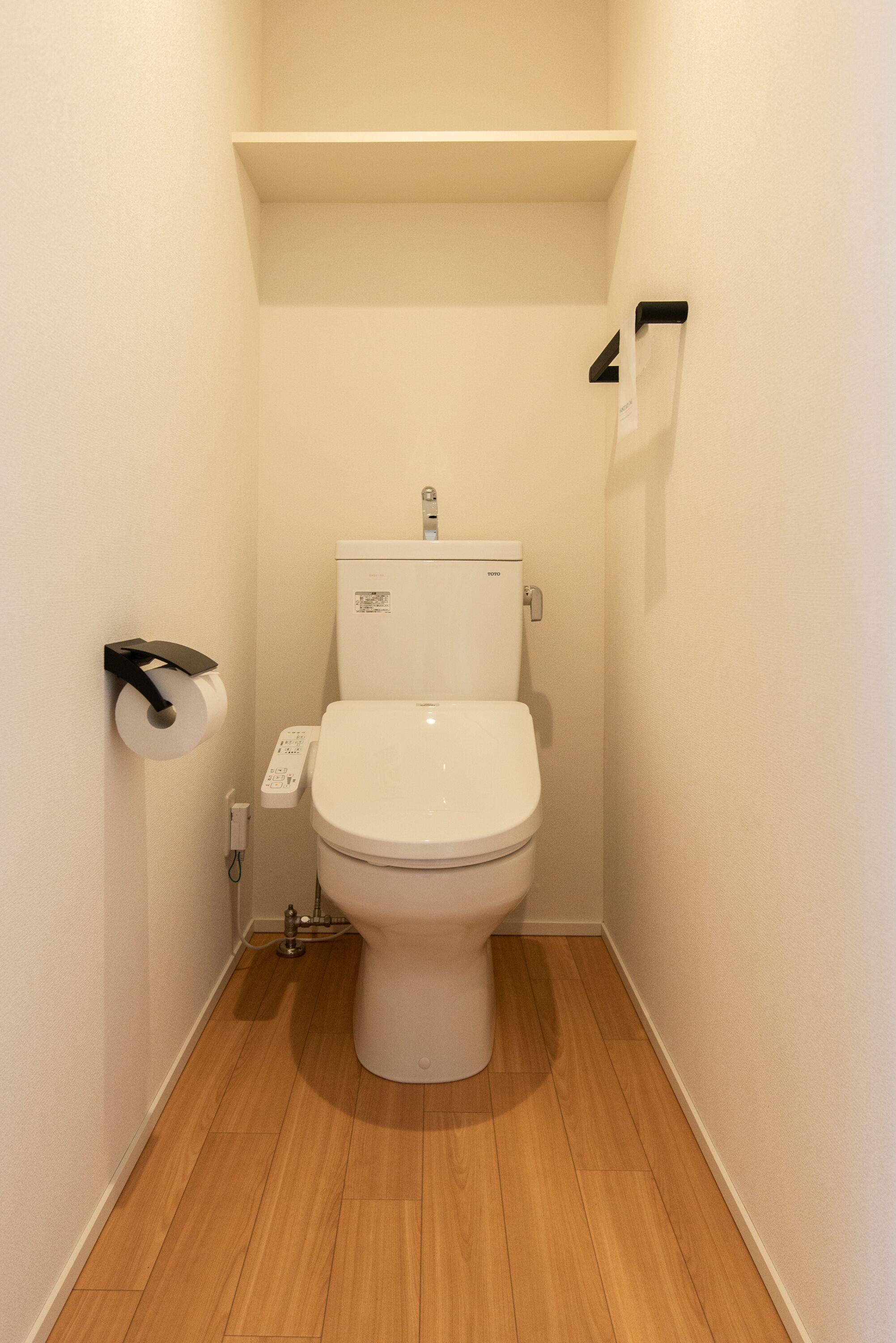 A small, narrow bathroom with a toilet, a toilet paper holder, and a shelf above the toilet.