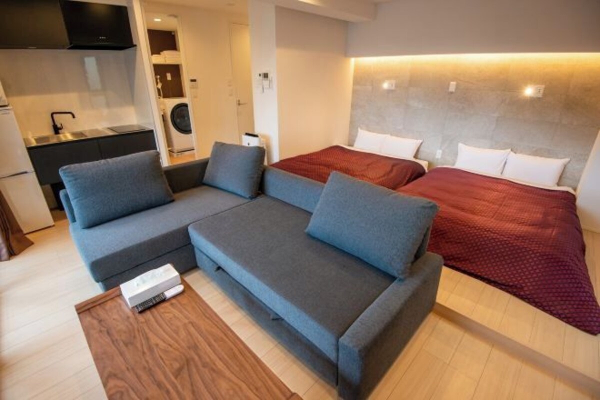 A modern hotel room with a sofa, two beds, a small kitchen area, and a washing machine.