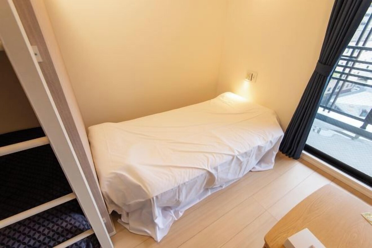 A small, single-bed room with a window, a bedside table, and a wooden floor.