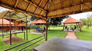 An outdoor dining area with tables and chairs under a thatched roof, surrounded by greenery and a building with a red roof. - Avadale Masinagudi - Stag Groups Not Allowed  (Ootacamund)