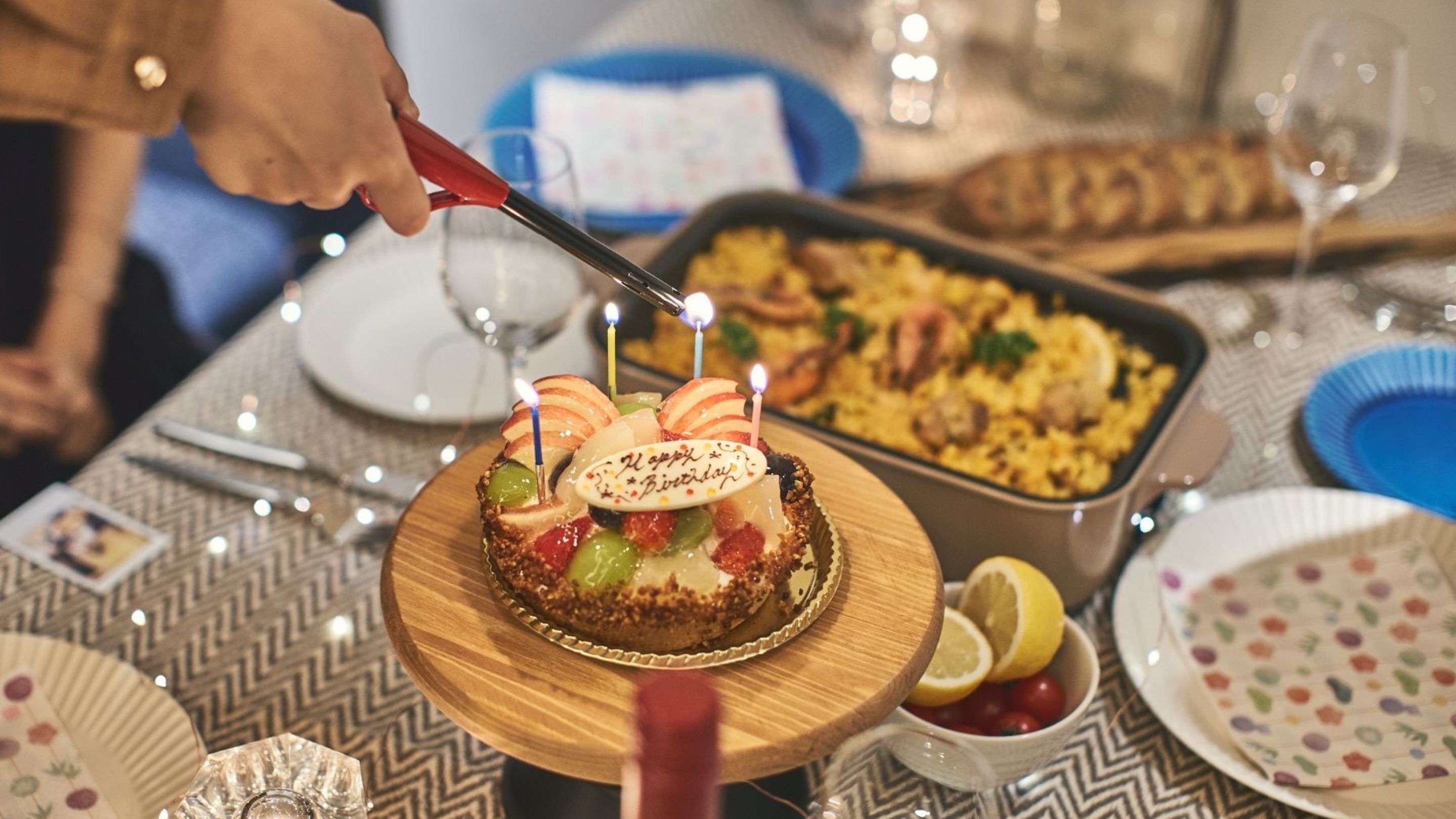 A birthday cake with candles on a wooden plate, surrounded by various dishes and a wine glass on a table.