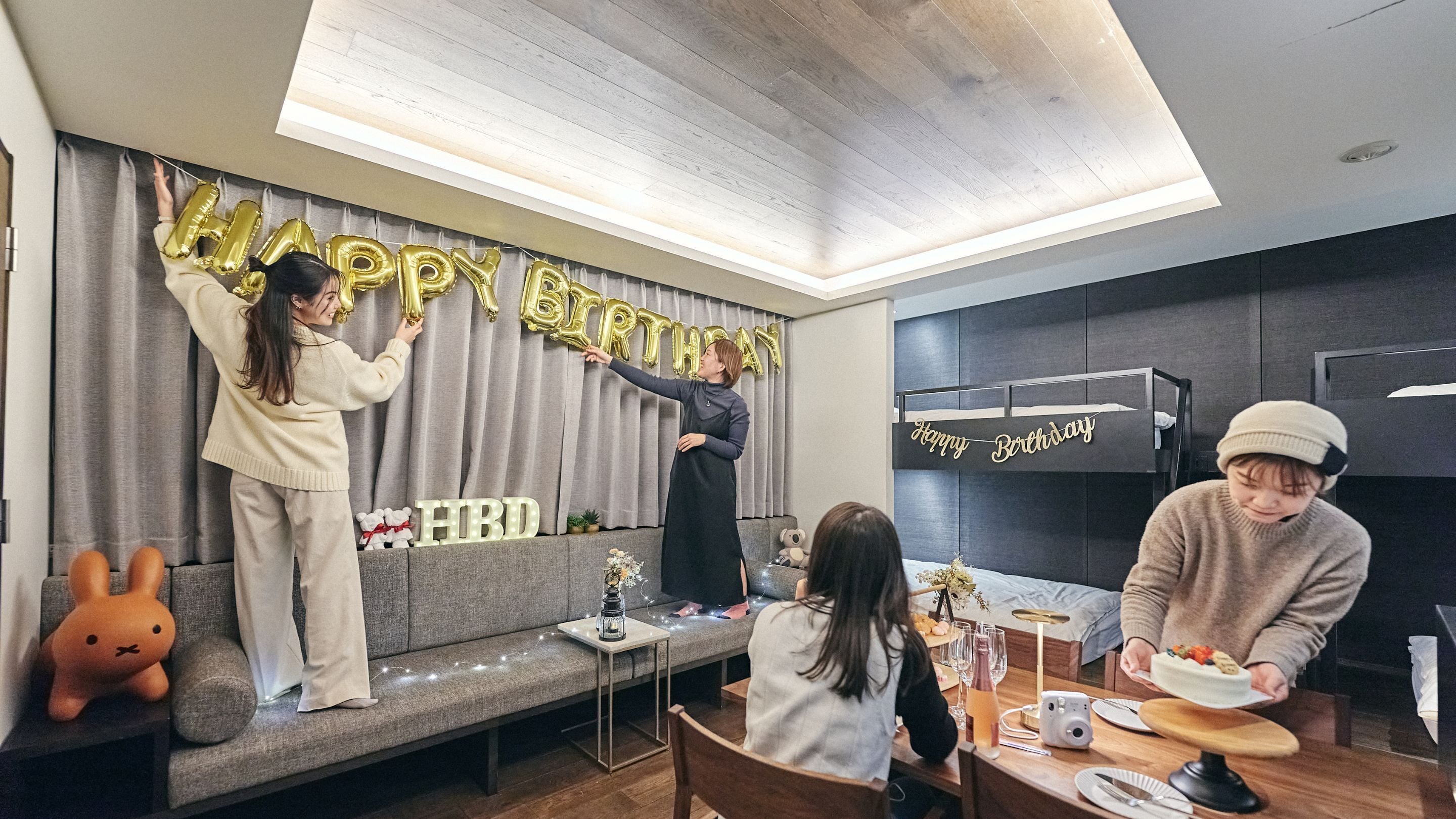 A modern living room with a grey sofa, a dining table with a cake, and a 'Happy Birthday' balloon decoration.