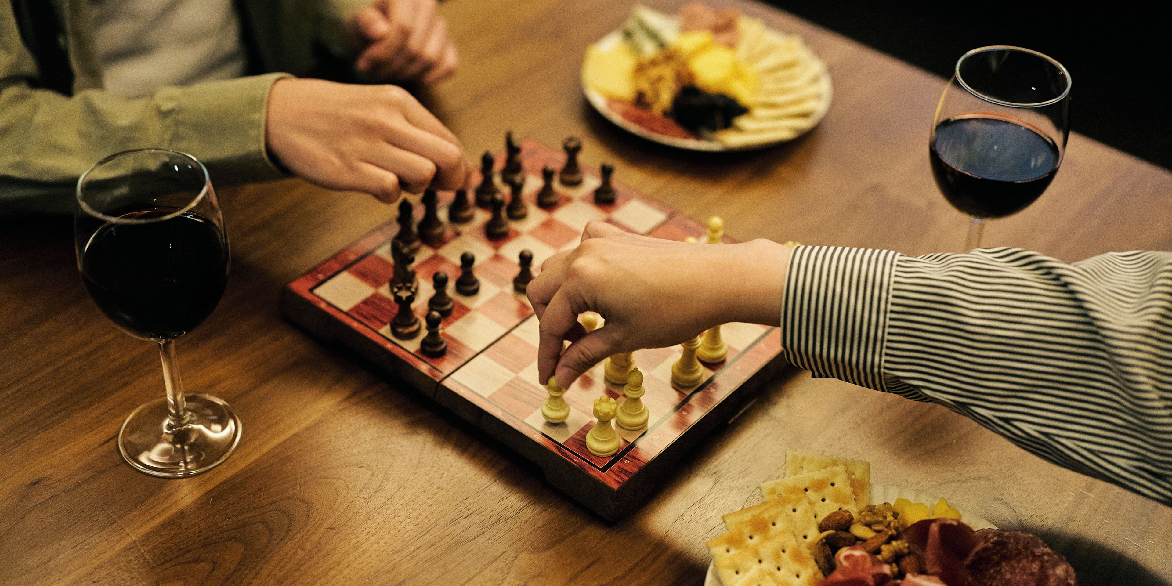 Un jeu d’échecs sur une table en bois, avec deux verres de vin rouge et une assiette de nourriture.