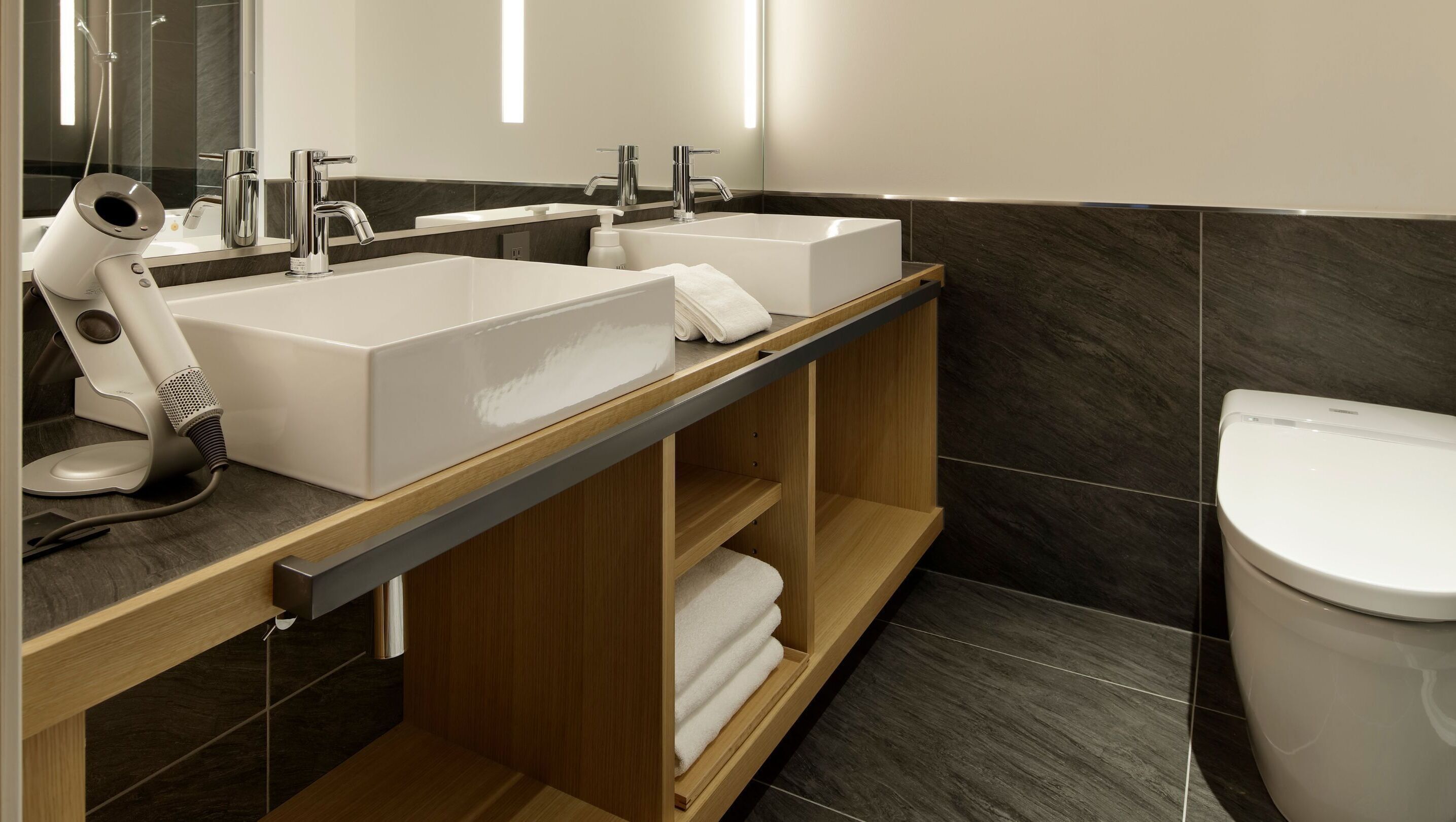 A modern bathroom with two sinks, a hairdryer, and a toilet.