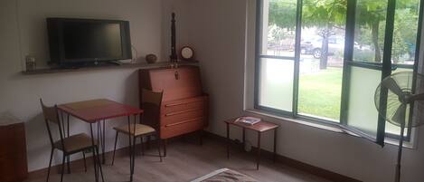 A room with a TV, a guitar, a dresser, a table with chairs, a small fan, and a window with a view of a garden.