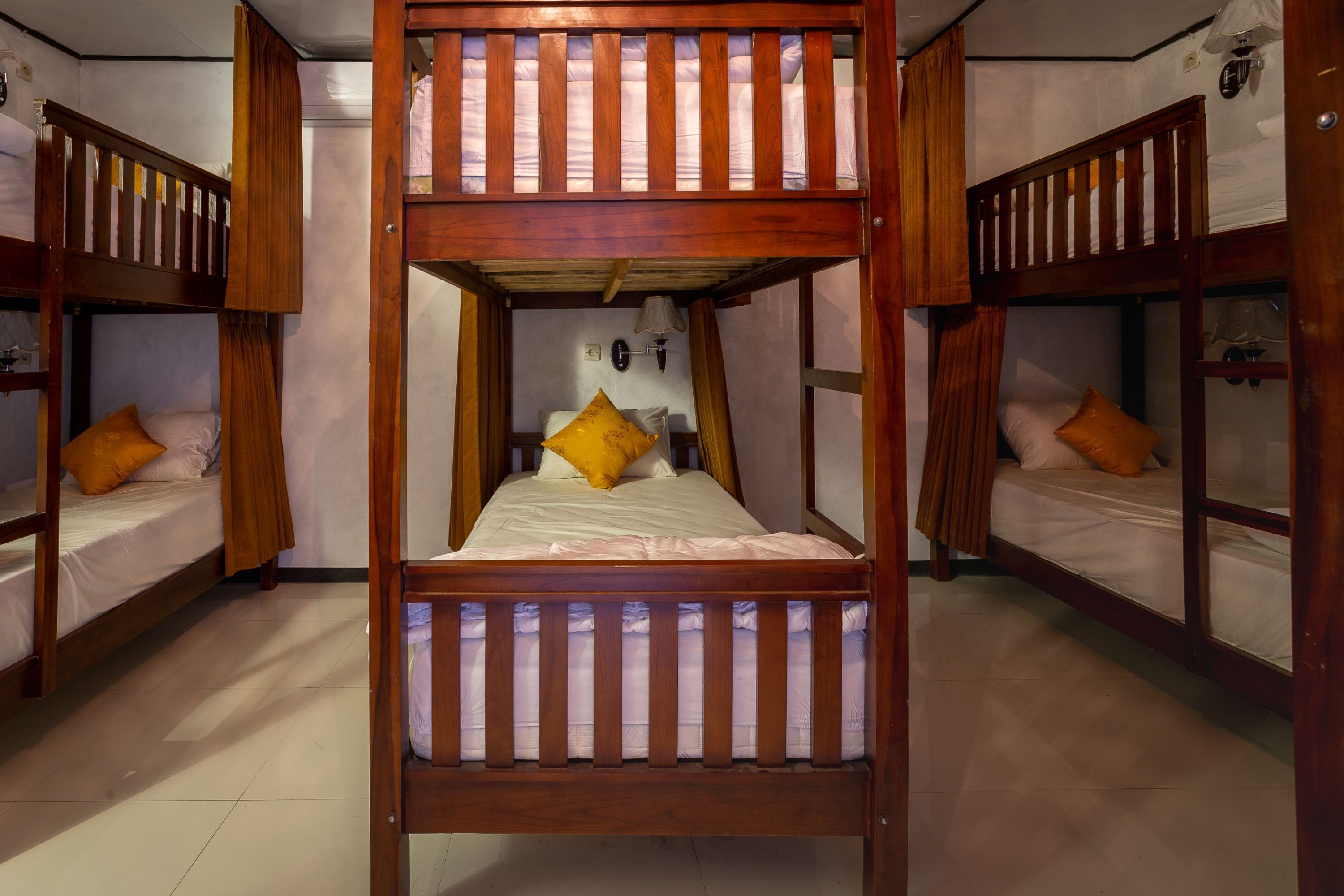 A room with three bunk beds, each with a wooden frame and white bedding. There is a wall-mounted lamp above the central bed.