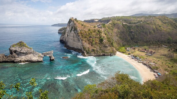 A coastal landscape with a prominent rock formation, clear turquoise waters, and a beach with structures. - Penida Krusty Hill (Penida Island)