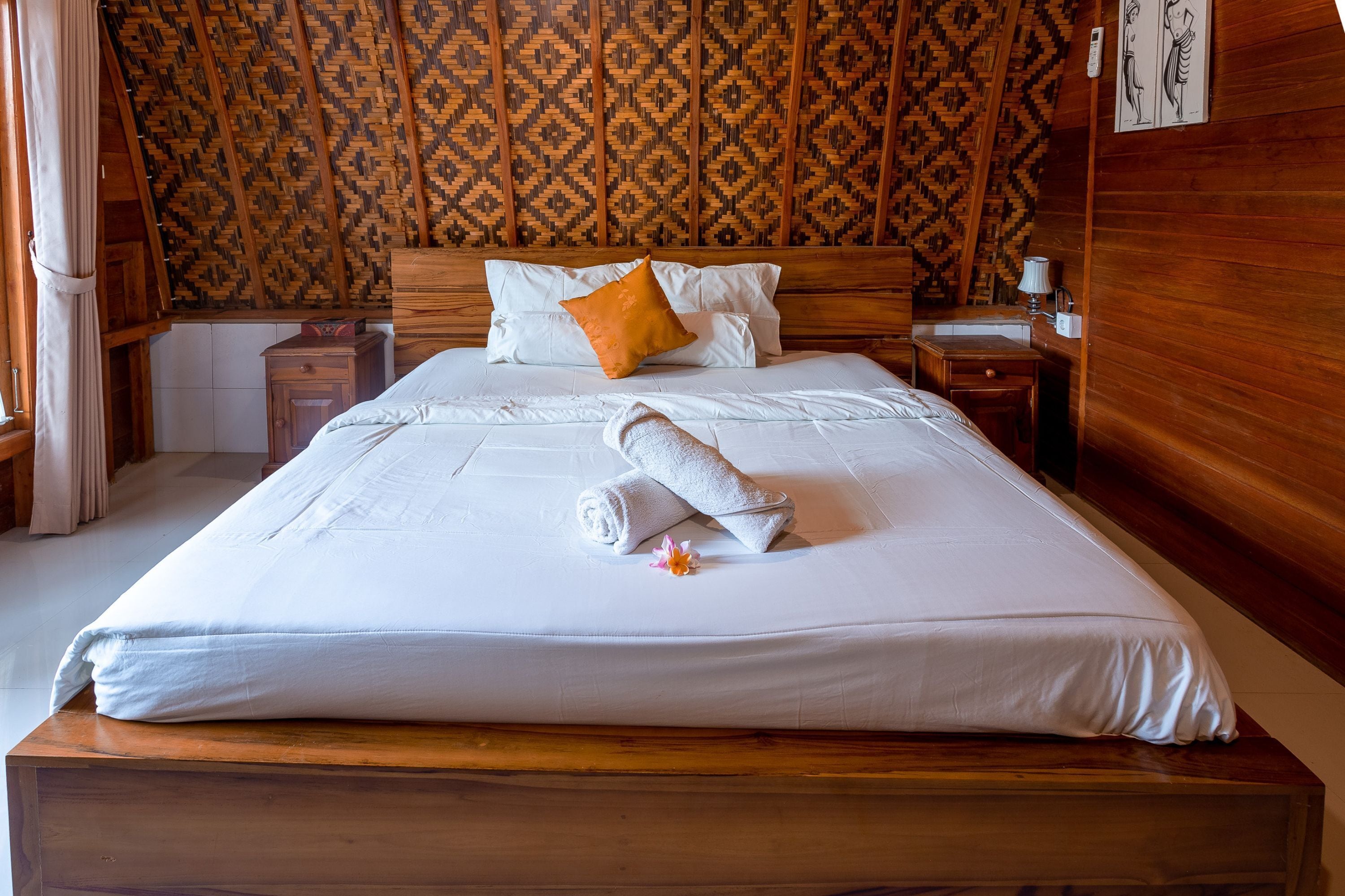 A neatly made bed with white linens and a rolled towel, a wooden headboard, and a patterned wall.