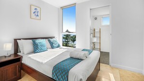 A modern hotel room with a large bed, a nightstand, a bedside lamp, a window with a view of palm trees, and a door leading to a bathroom. - Lakes Hotel Serviced apartments (Toukley)