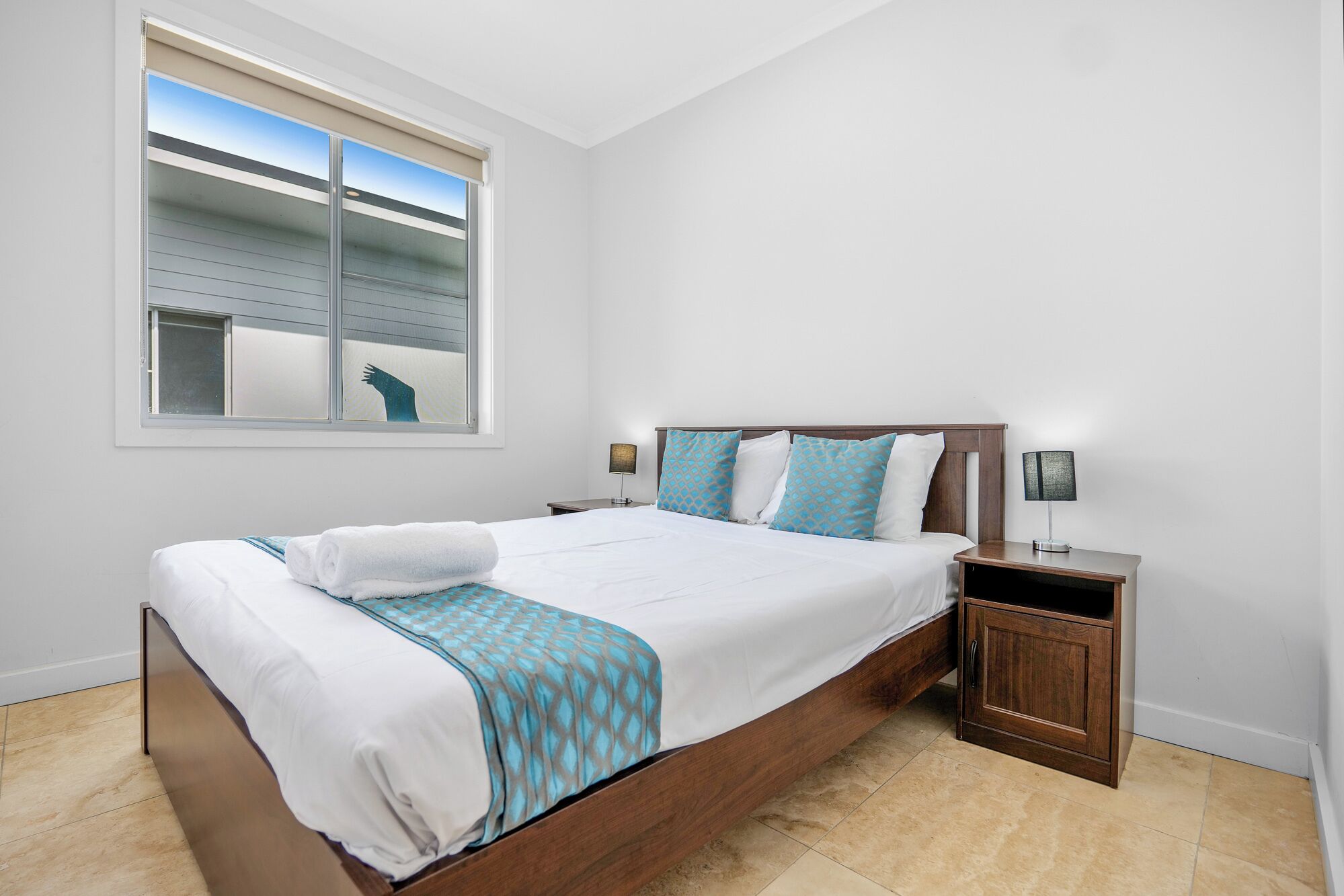 A bedroom with a large bed, a nightstand, a lamp, and a window with blinds.