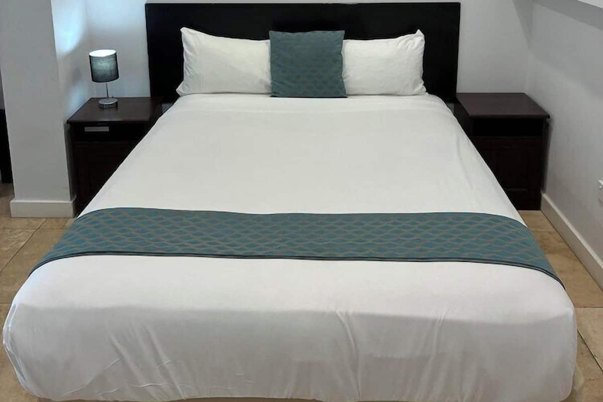 A neatly made bed with a white and teal bedspread, a dark headboard, and a single bedside lamp.