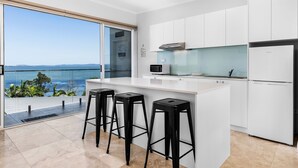 A modern kitchen with a large island, white cabinetry, and a view of the ocean. - Lakes Hotel Serviced apartments (Toukley)