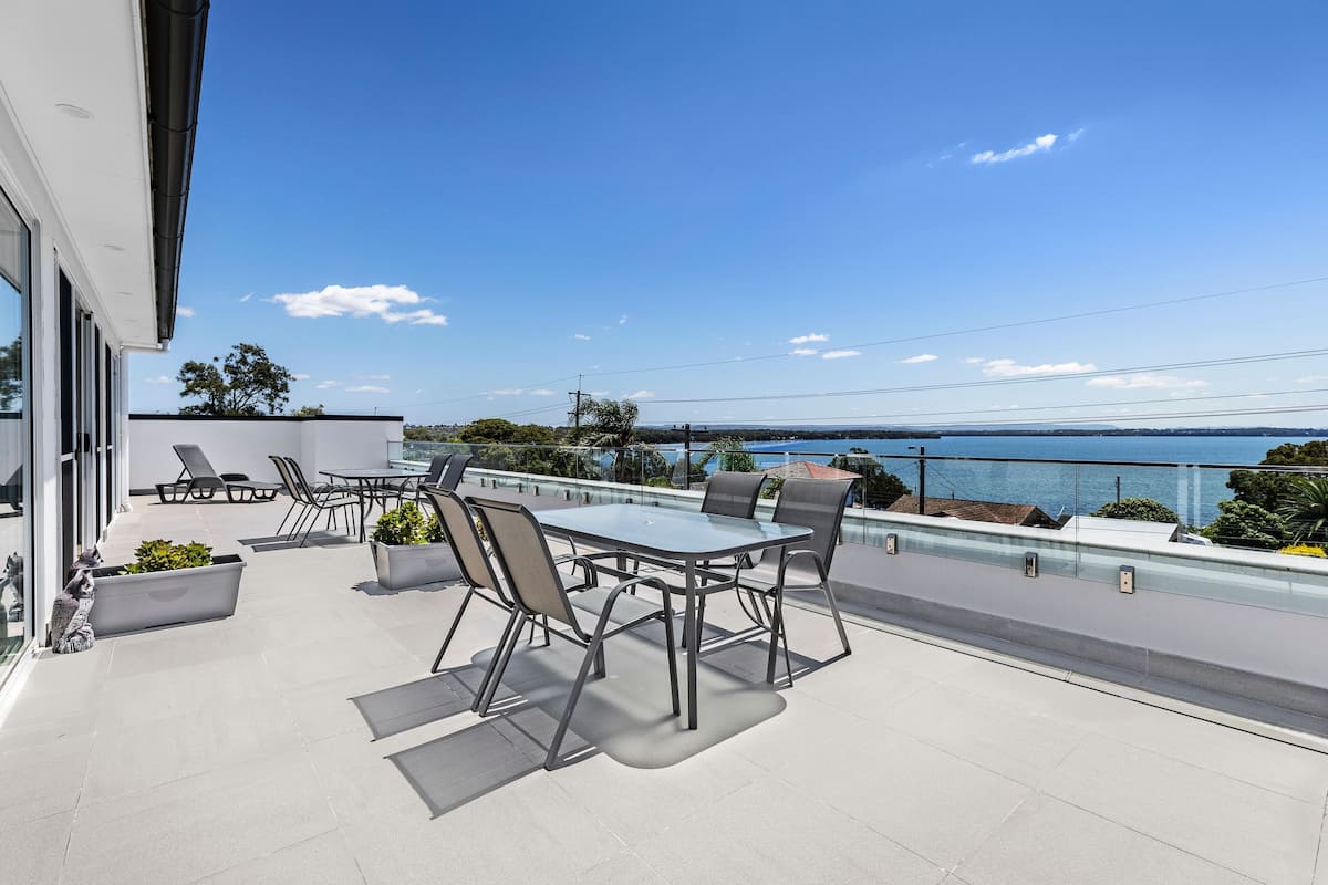 Eine moderne Außenpatio mit Glastisch und -stühlen, umgeben von Topfpflanzen, mit Blick auf eine malerische Seelandschaft.