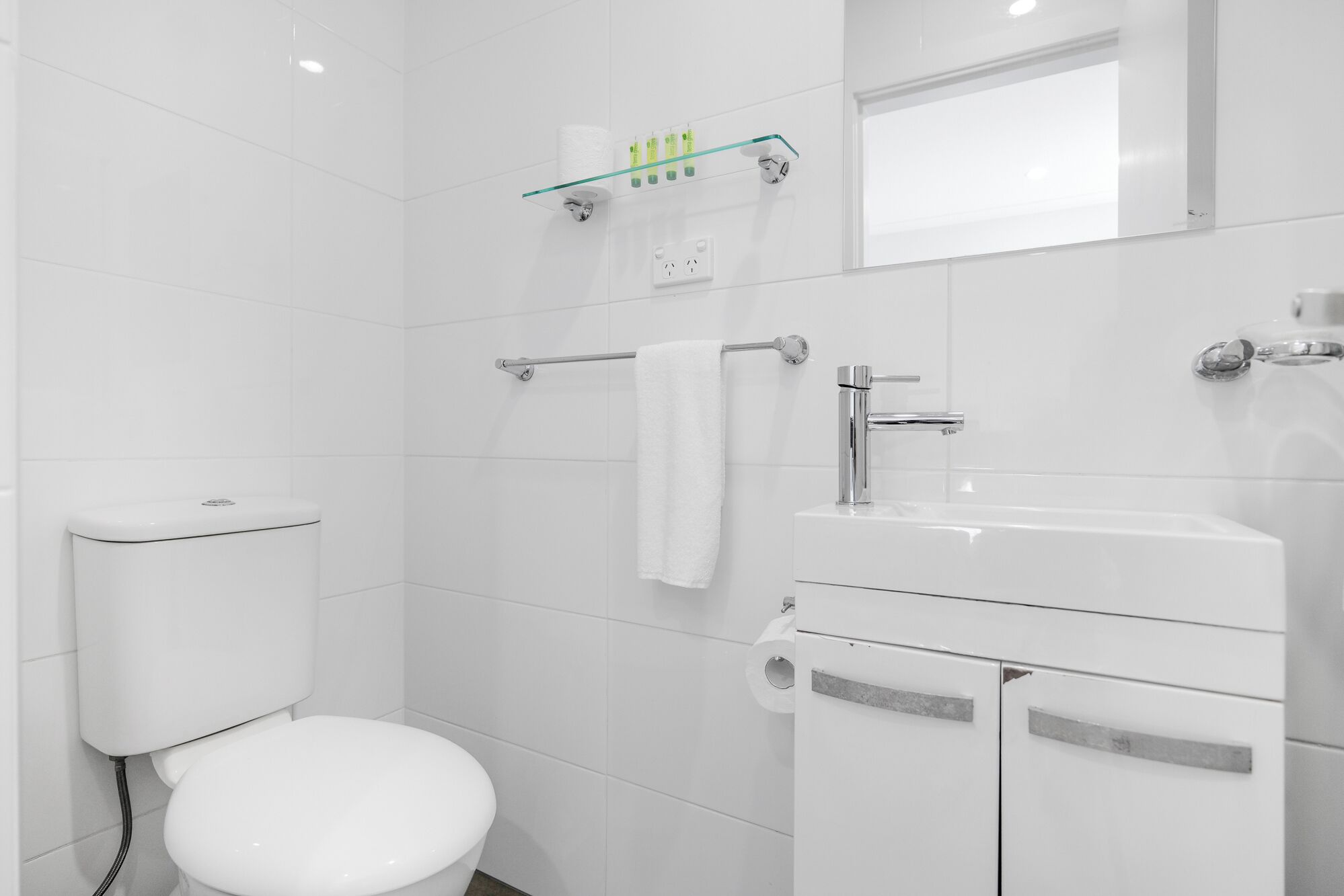 A modern bathroom with a toilet, a glass shelf with toiletries, a towel rack, and a sink.