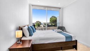 A bedroom with a large window, a bed with white bedding, a bedside table with a lamp, and a view of the outside.