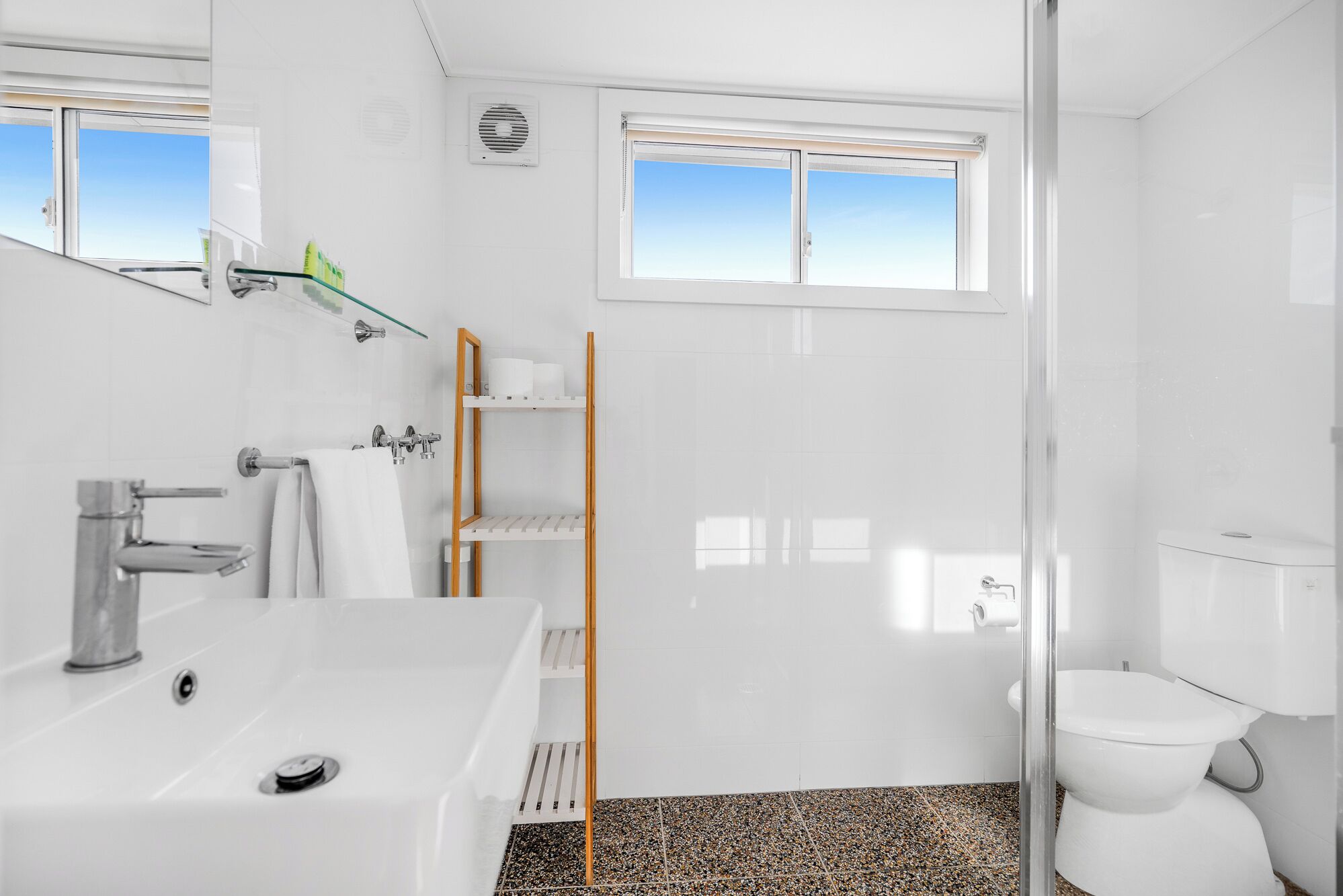 A modern bathroom with a white sink, mirror, and toilet. There is a glass shower enclosure and a window above the toilet.