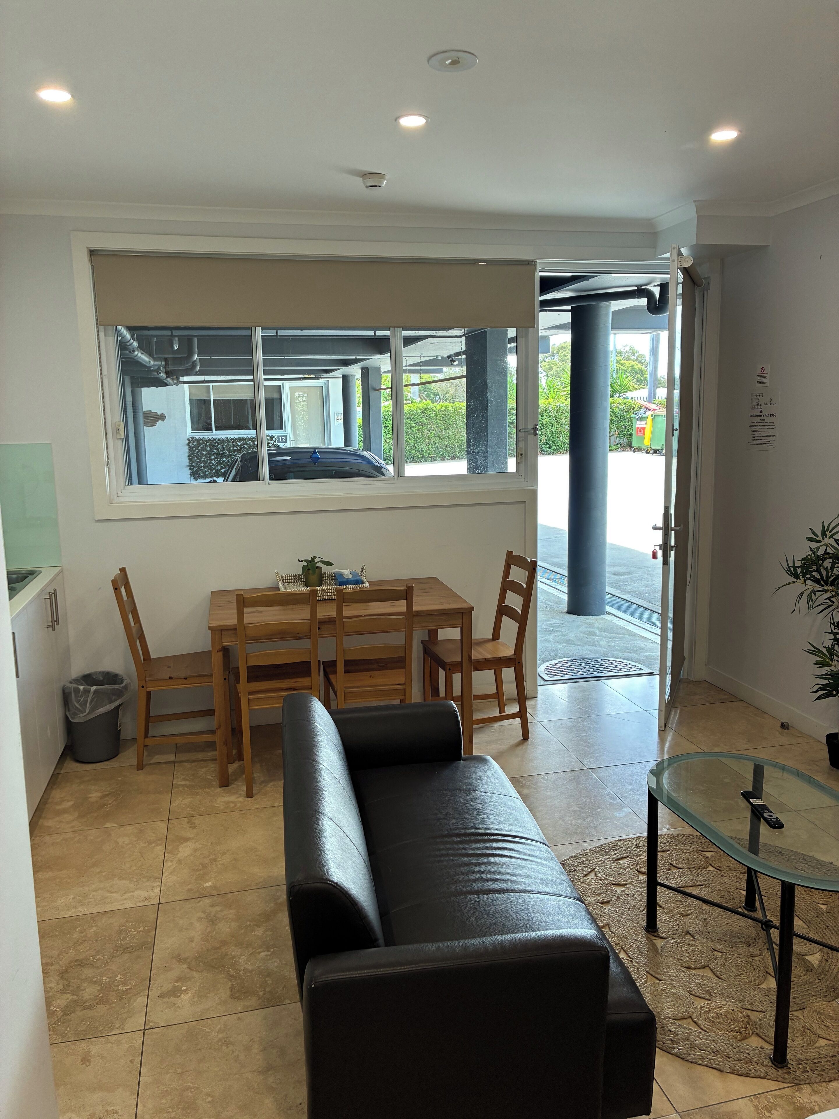 A room with a dining table, chairs, a black leather sofa, a glass coffee table, and a window with a view of the outside.