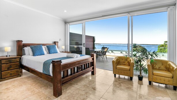 A bedroom with a large bed, a nightstand, a bedside table, a window with a view of the sea, and two chairs. - Lakes Hotel Serviced apartments (Toukley)