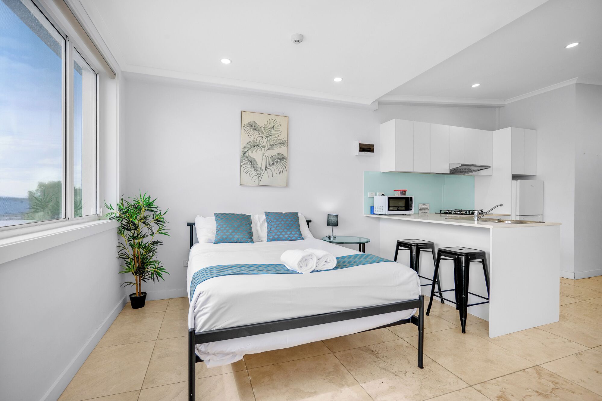 A modern hotel room with a bed, a small kitchen, a dining area, and a view of the sea.