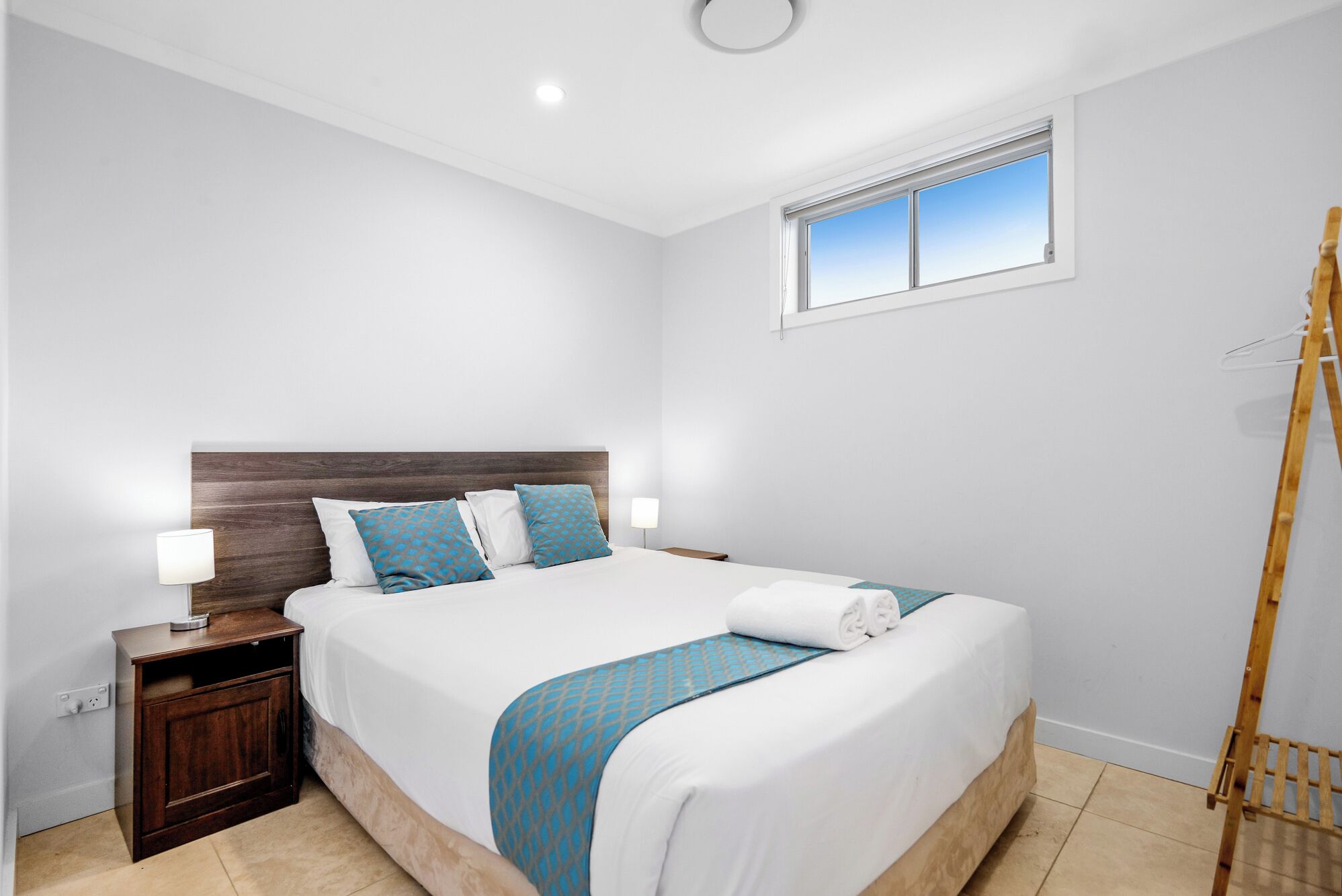 A bedroom with a wooden headboard, a bed with white bedding and blue accents, a bedside table with a lamp, and a window with a view.