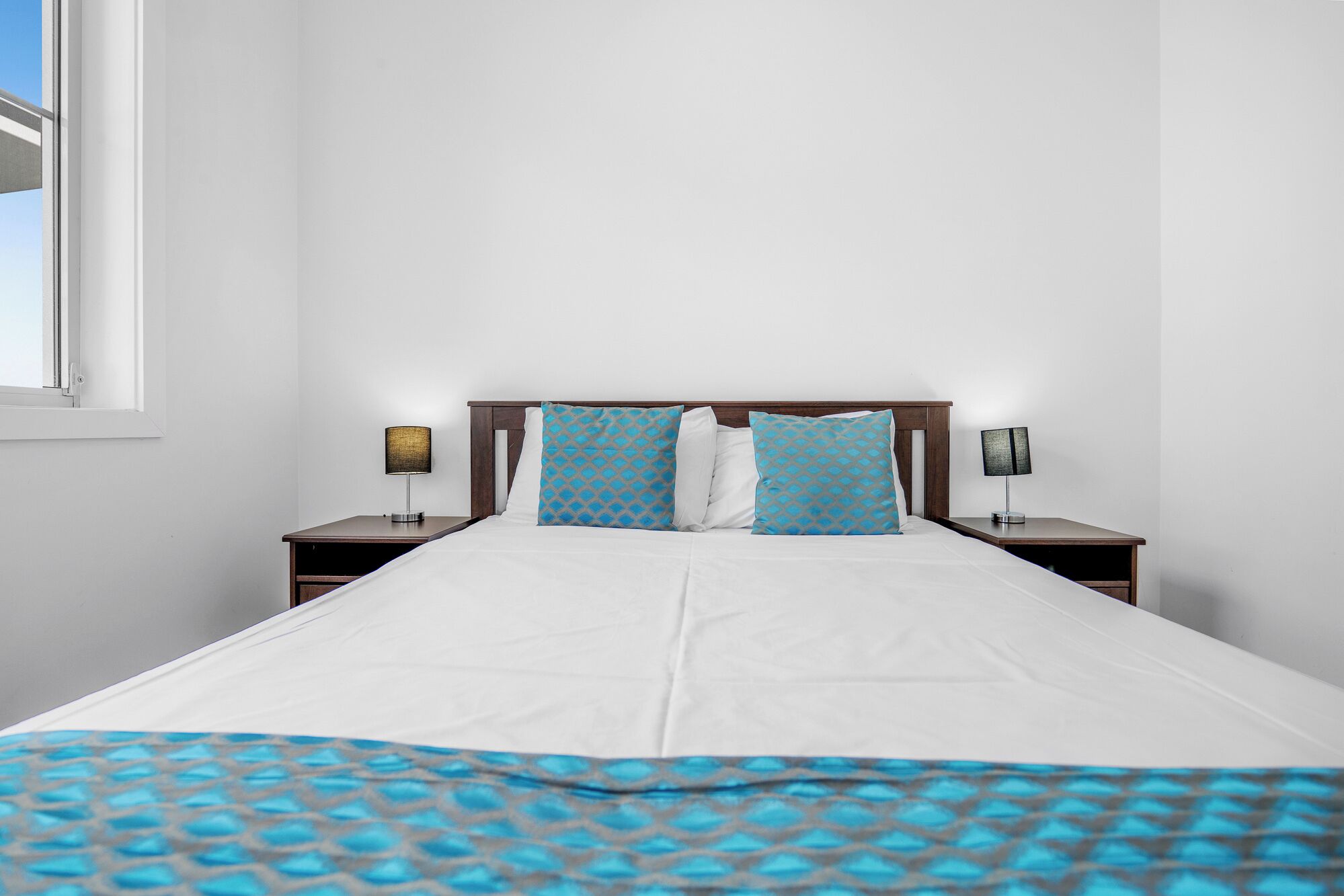 A neatly made bed with a white sheet and a blue patterned throw blanket. There are two blue patterned pillows on the bed. The room has white walls and a window on the left side.
