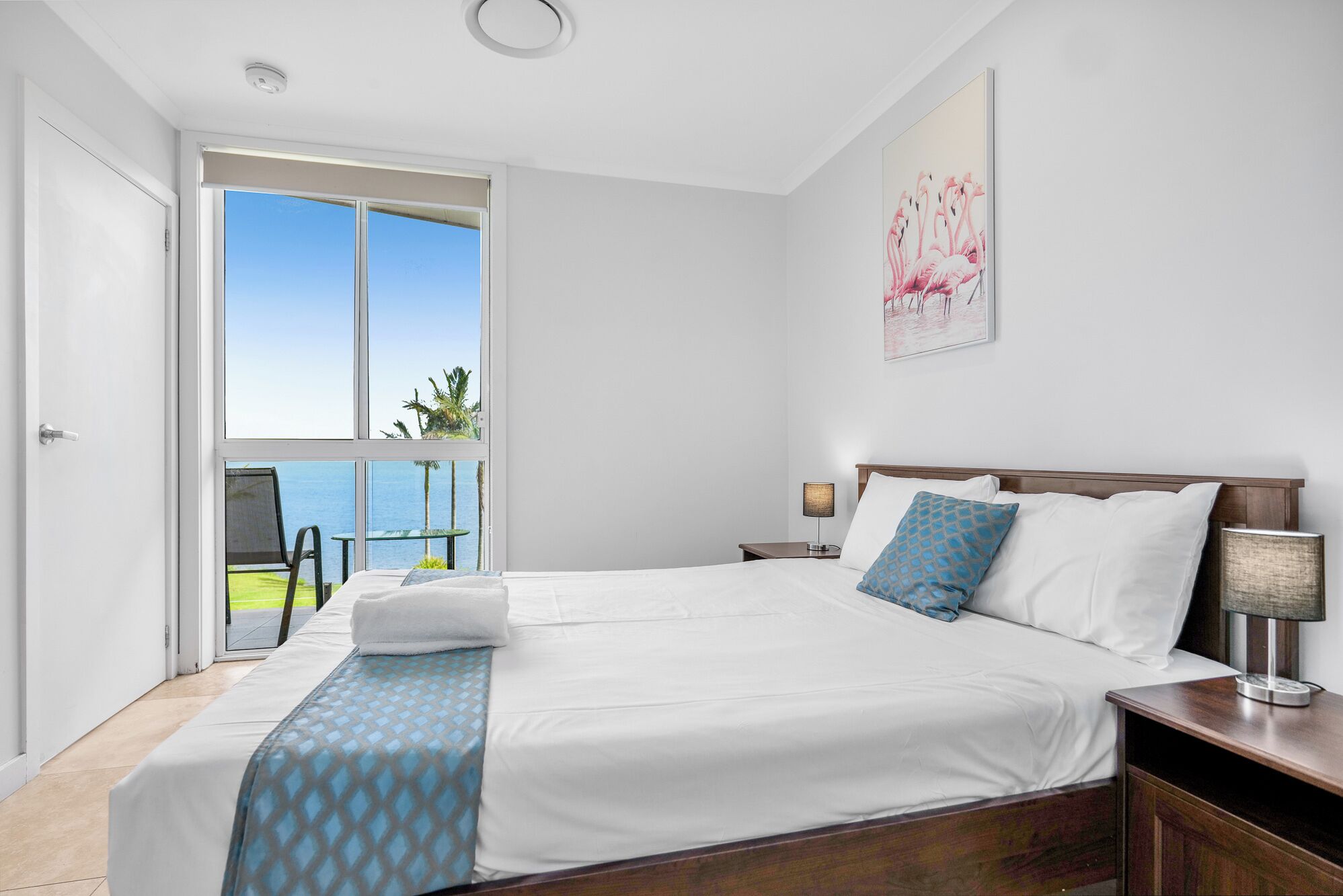 Quarto de hotel com uma cama grande, uma mesa de cabeceira, uma luminária e vista para o oceano através de uma janela ampla.