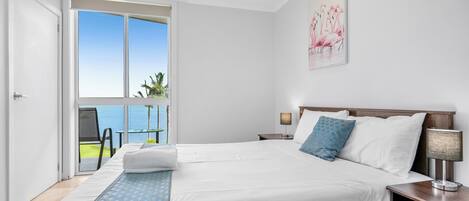 Quarto de hotel com uma cama grande, uma mesa de cabeceira, uma luminária e vista para o oceano através de uma janela ampla.