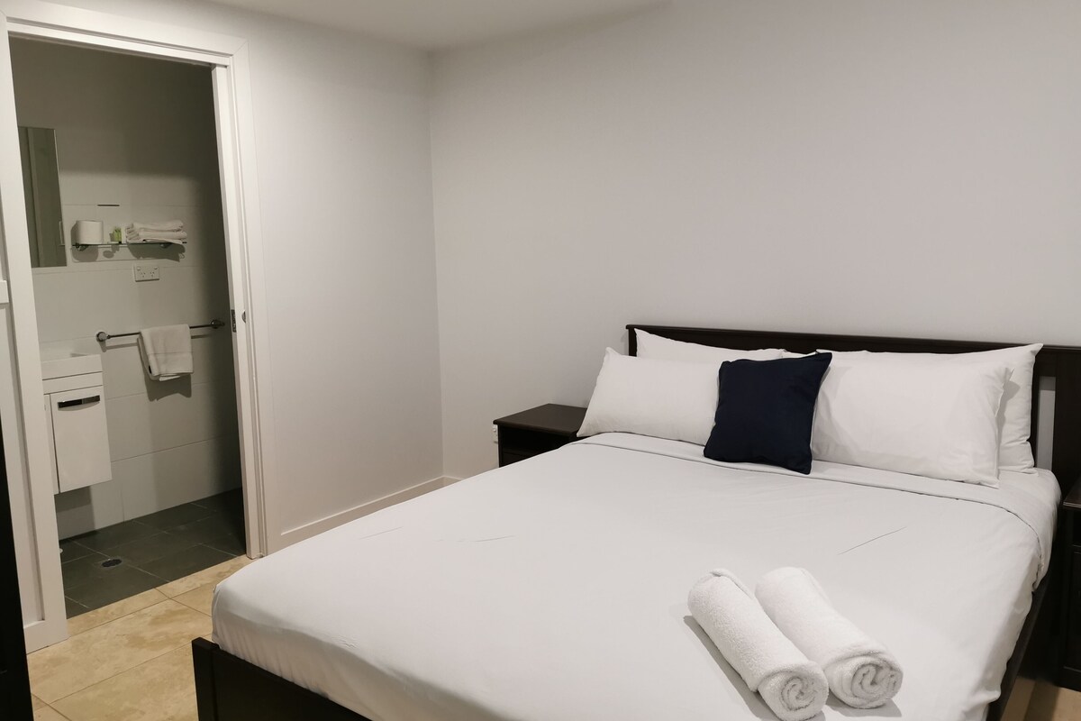 Habitación de hotel limpia y moderna con una cama bien hecha, una mesita de noche y un baño visible a través de una puerta abierta.