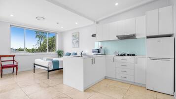 Una cocina moderna con armarios blancos, un microondas, una estufa y un refrigerador. Hay una ventana con vistas a árboles y una silla roja en el comedor.