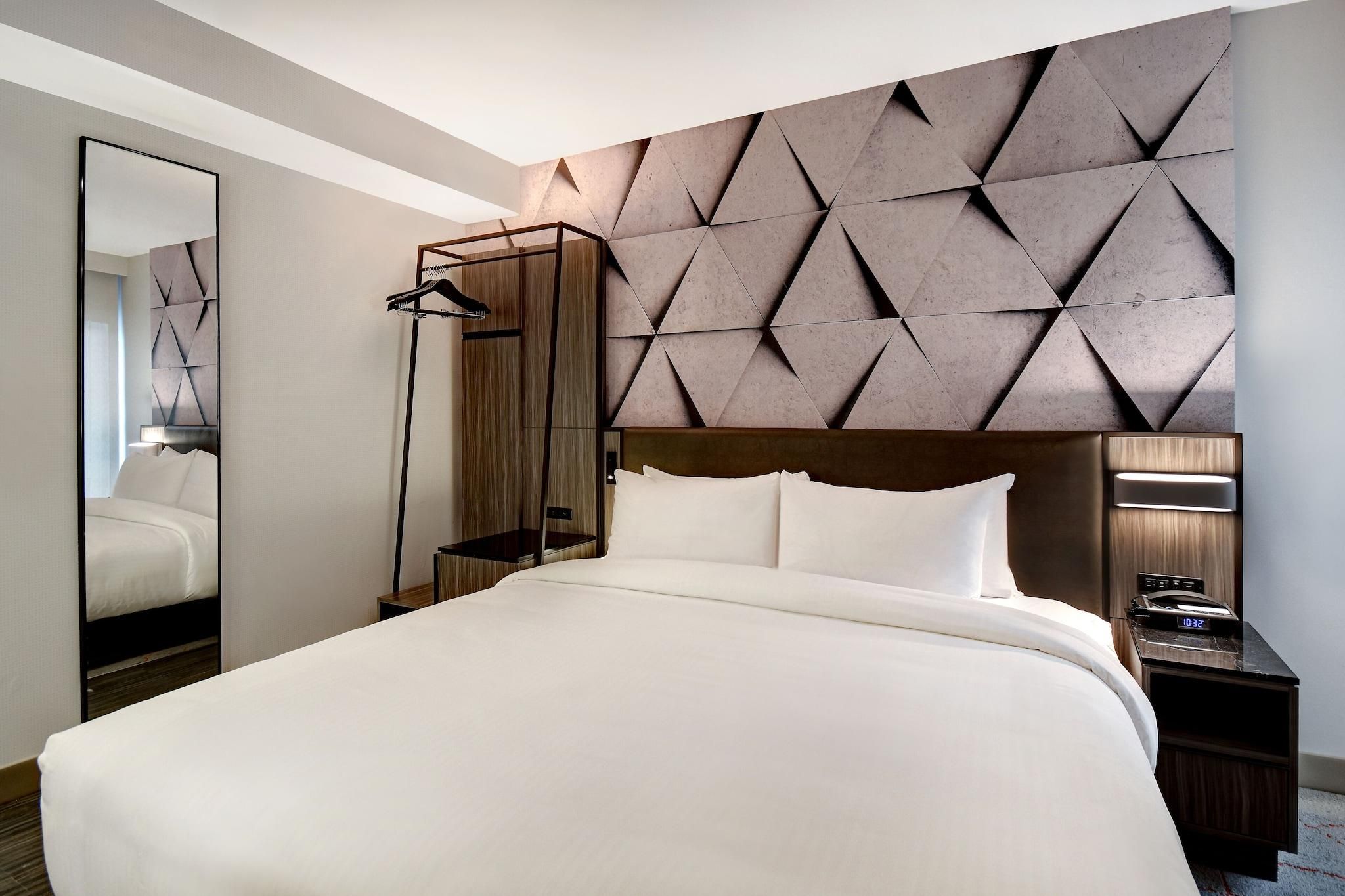 A hotel room with a large bed, a mirror, a wooden headboard, a nightstand with a clock, and a geometric-patterned wall.