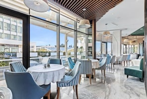 A modern restaurant with round tables and striped chairs, large windows, and a view of buildings and water. - Barbeta Accommodation (Budva)