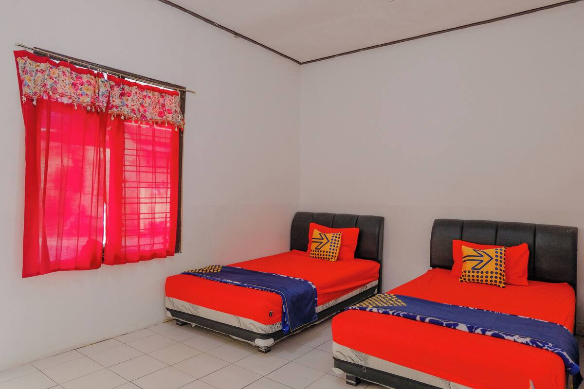 Ein Zimmer mit zwei Betten, jeweils mit roter Bettwäsche und gemusterten Kissen, einem Fenster mit roten Vorhängen und einem gefliesten Boden.