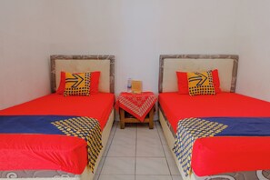 A room with two beds, each with red bedding and patterned pillows. A small wooden table with a red cloth and a bottle of water is between the beds.