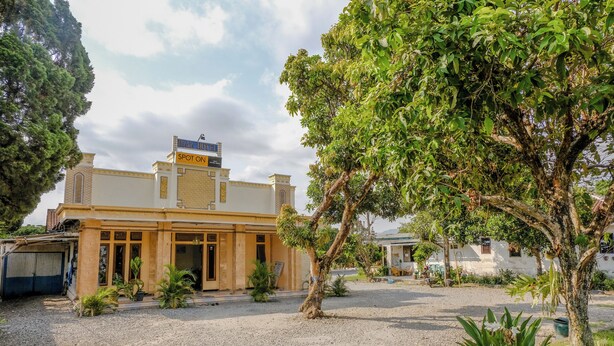 A building with a sign reading "LATE HOTEL SPOT ON." It features a courtyard with gravel, potted plants, and a large tree. - Spot On 1847 Hotel Maya (Beru)